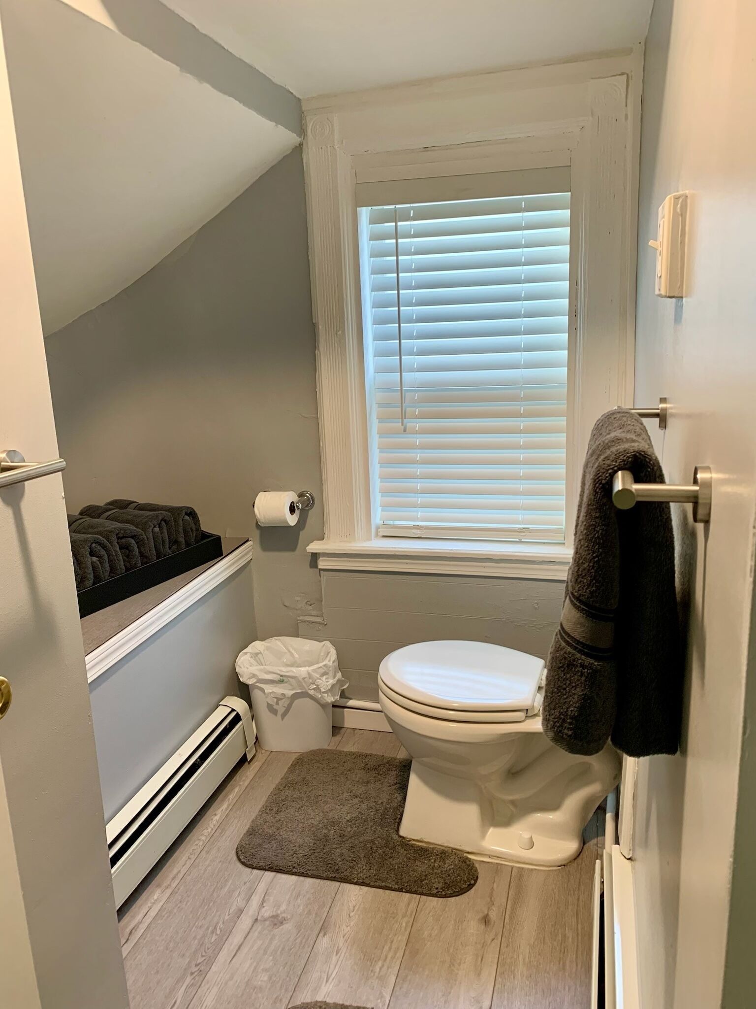 Une petite salle de bain comprenant des toilettes, une petite fenêtre avec stores, un porte-serviettes et une bouche d’aération.