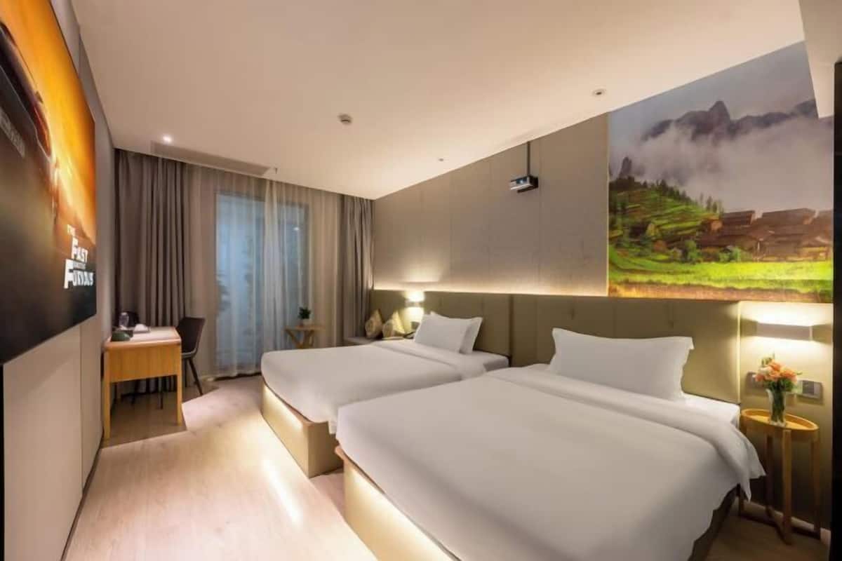 A hotel room with two beds, a desk, a chair, a window with curtains, and a painting on the wall.