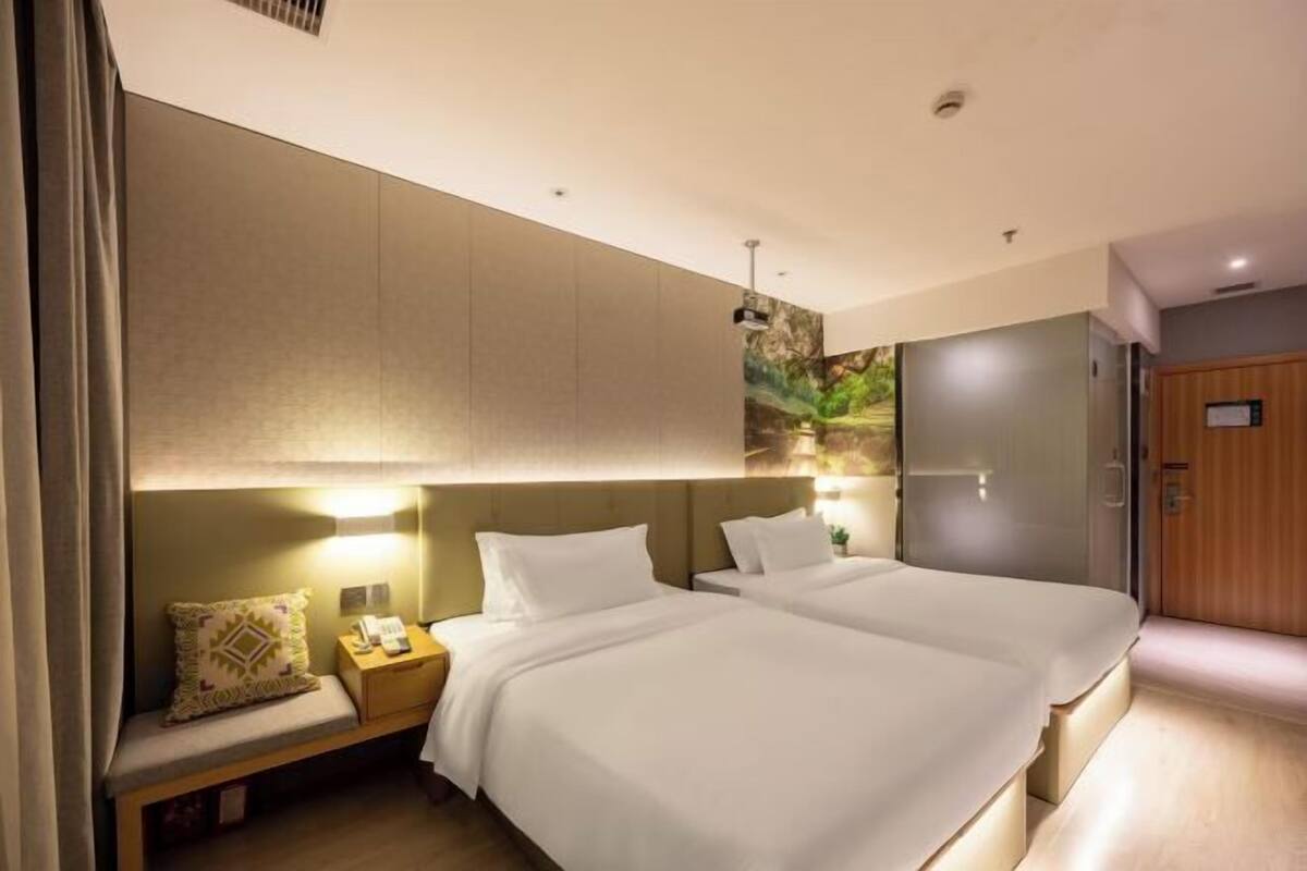 A hotel room with two beds, a bedside table with a lamp, a wall-mounted artwork, and a door.