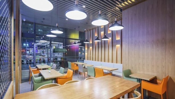 Un espace de restauration moderne avec des tables et des chaises en bois, un éclairage suspendu et une vue sur le paysage urbain grâce à de grandes fenêtres.
