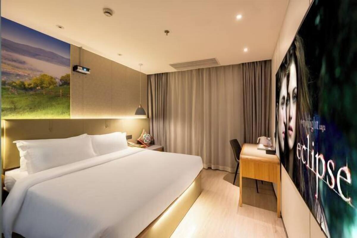 A hotel room with a large bed, a desk, a chair, a poster, and a window with curtains.
