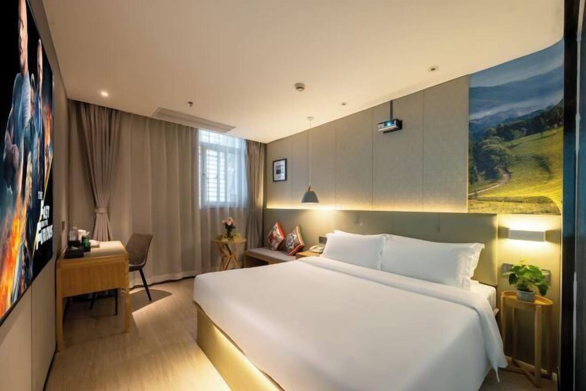 A hotel room with a large bed, a desk, a chair, a nightstand, a lamp, and a poster on the wall.