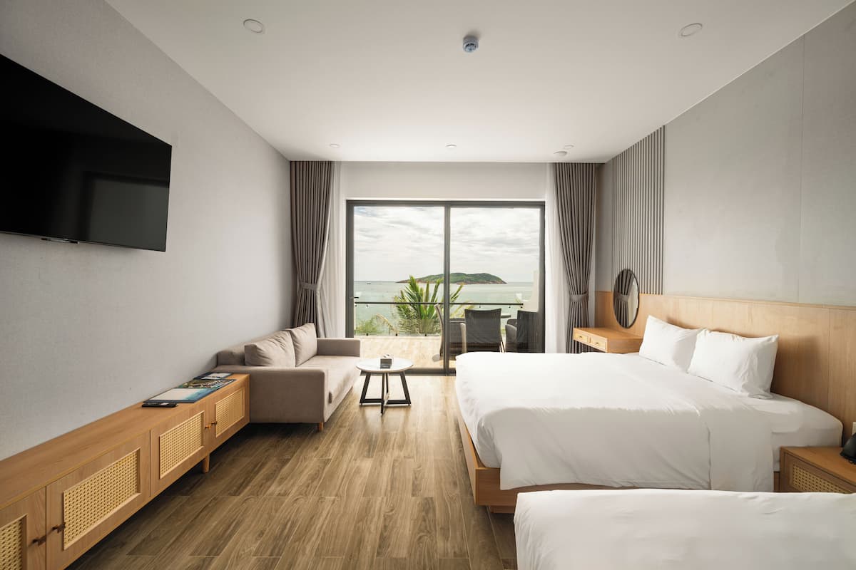A modern hotel room with a large bed, a sofa, a TV, and a balcony with a view of the sea.
