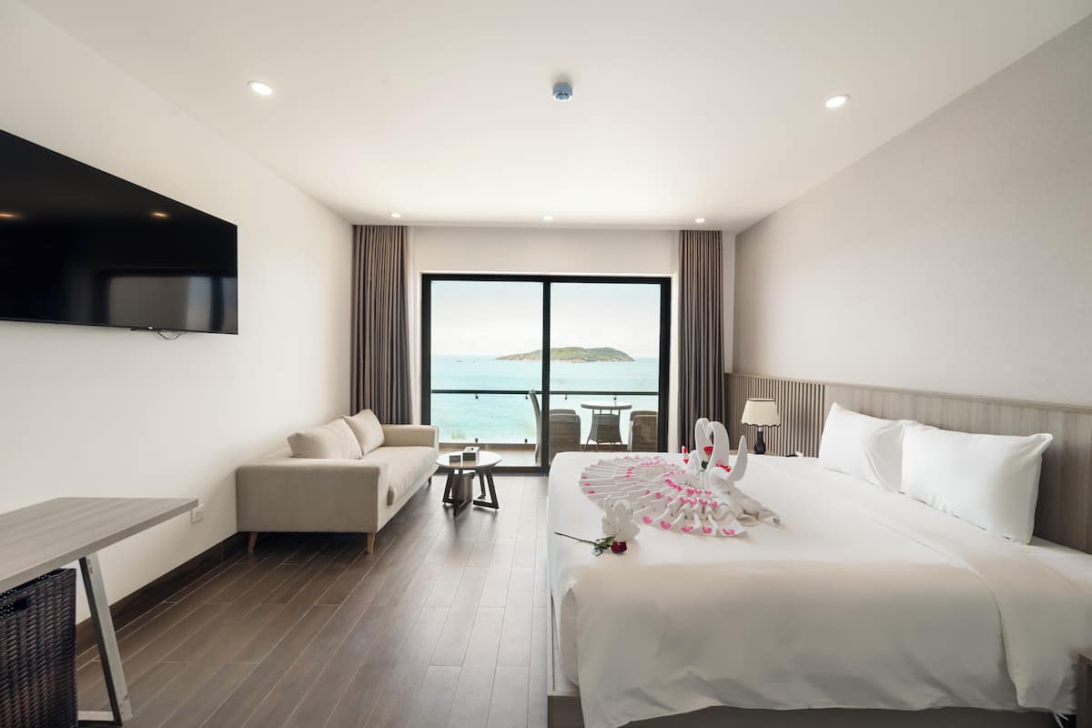 A hotel room with a large bed, a sofa, a small table, a TV mounted on the wall, and a view of the sea through a large window.