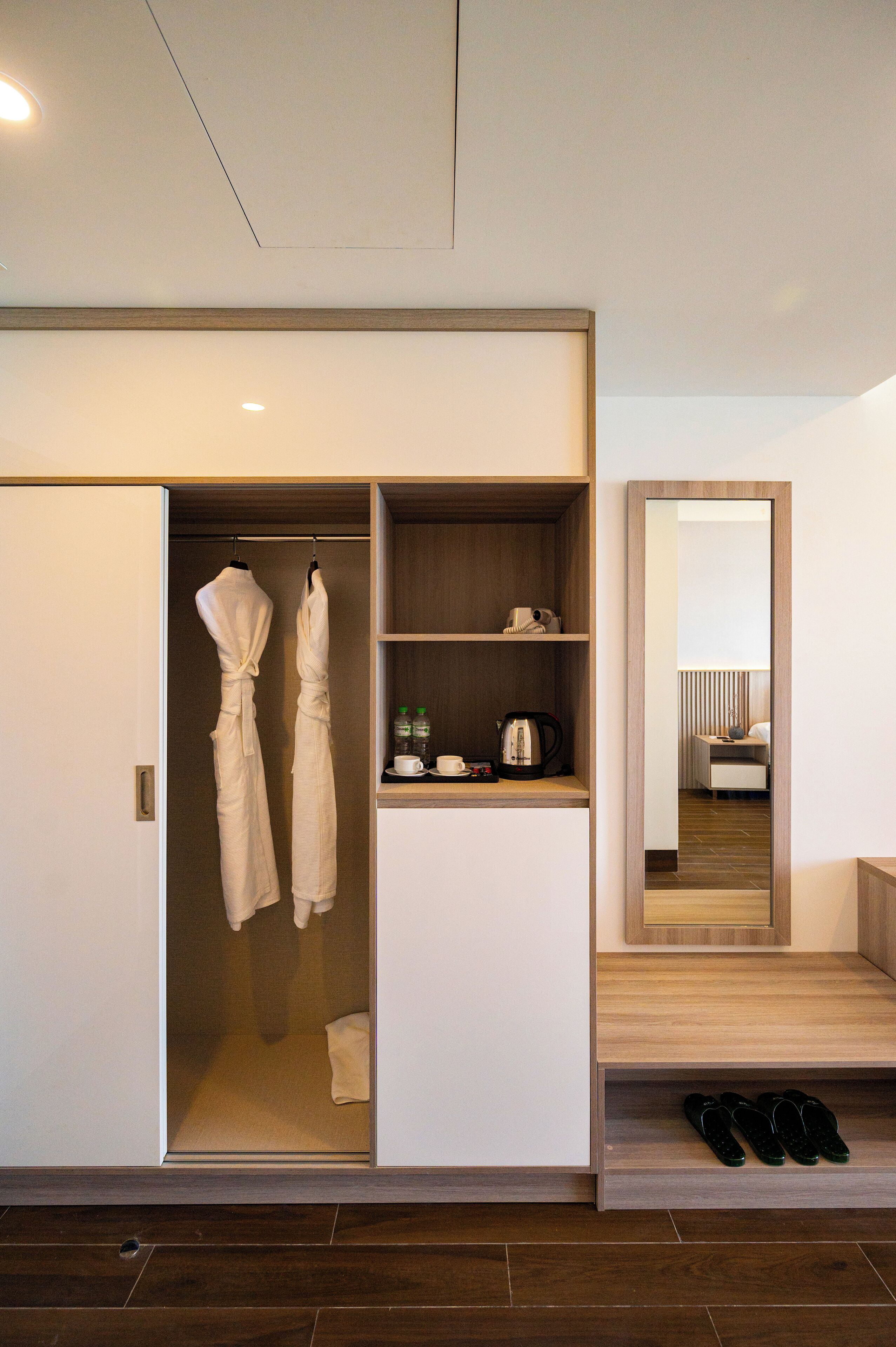 A hotel room with a walk-in closet containing robes, a coffee maker, and toiletries. There is a mirror reflecting the room and a pair of slippers on the floor.