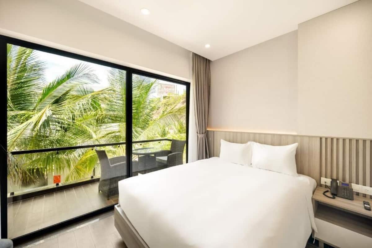 A hotel room with a large window offering a view of palm trees, a bed, a bedside table with a phone, and a chair by the window.
