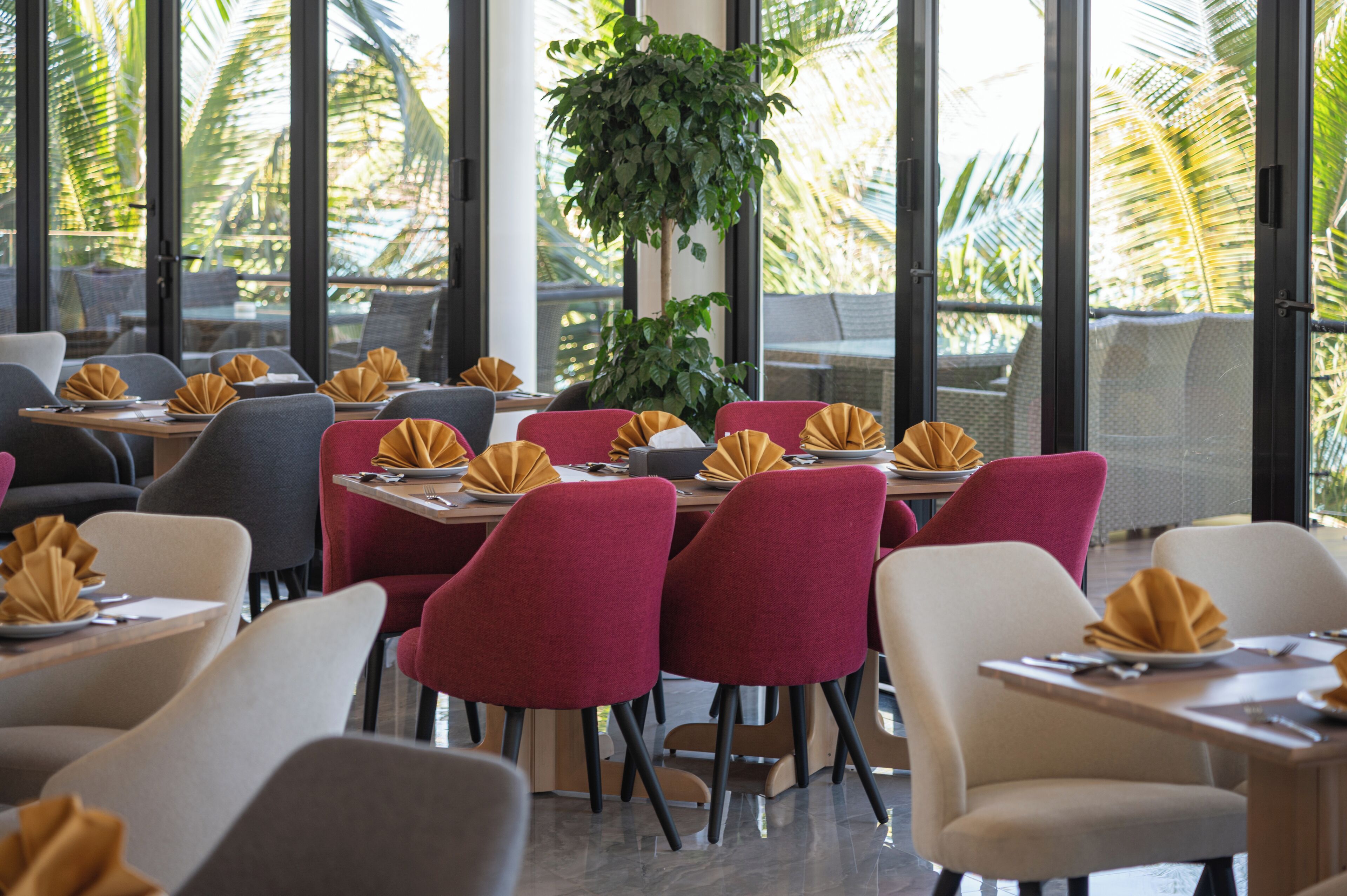 Un restaurant où les tables sont dressées pour le service, avec un mélange de chaises rouges et grises, et de grandes fenêtres offrant une vue sur les palmiers.