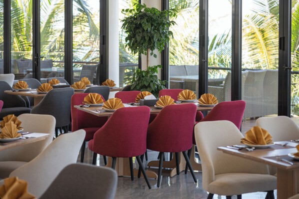 A restaurant with tables set for dining, featuring a mix of red and grey chairs, and large windows with a view of palm trees. - Quy Nhon Sea Resort (Quy Nhon Nam)