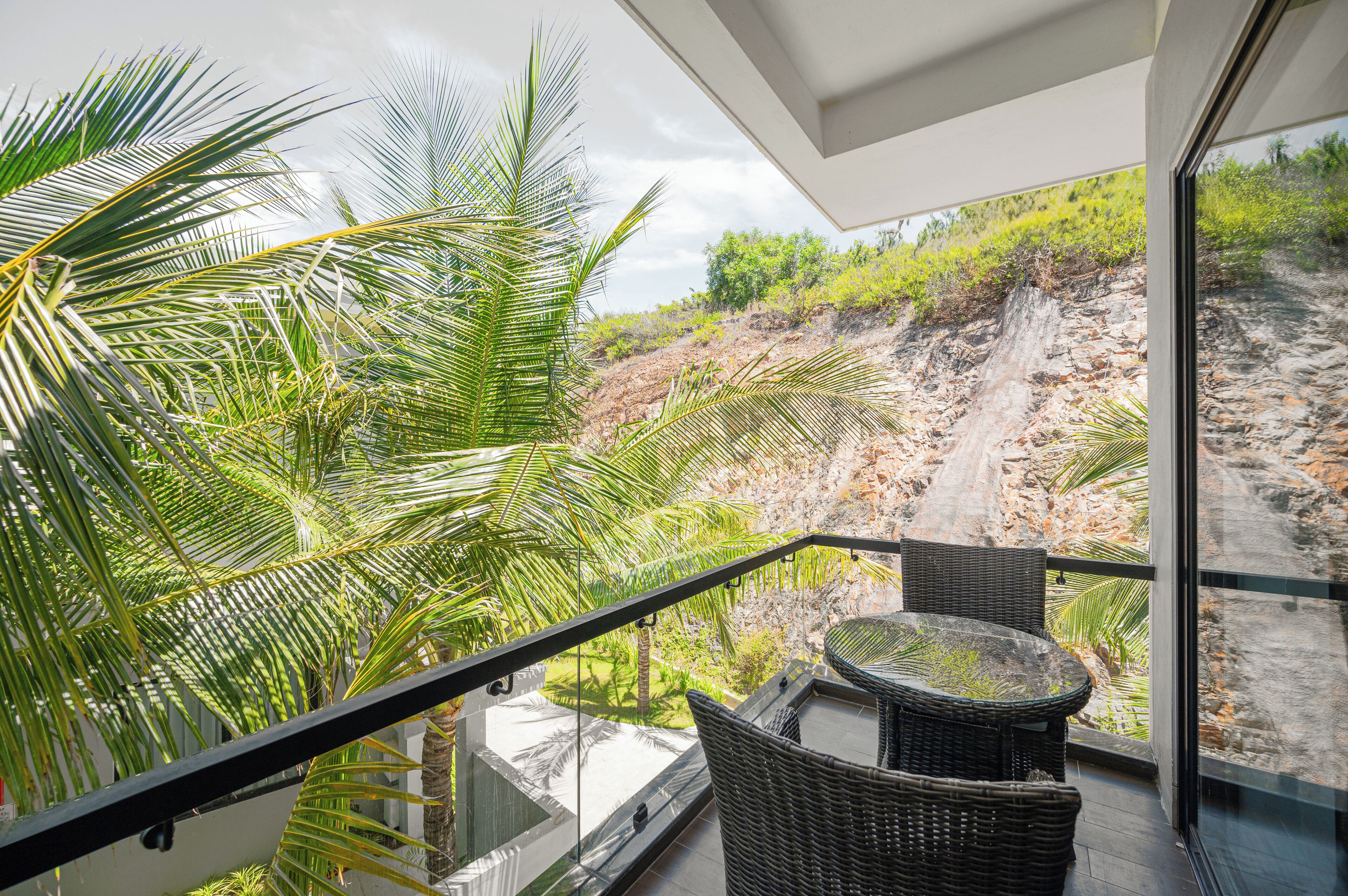 A balcony with wicker furniture overlooking a rocky hillside and palm trees.