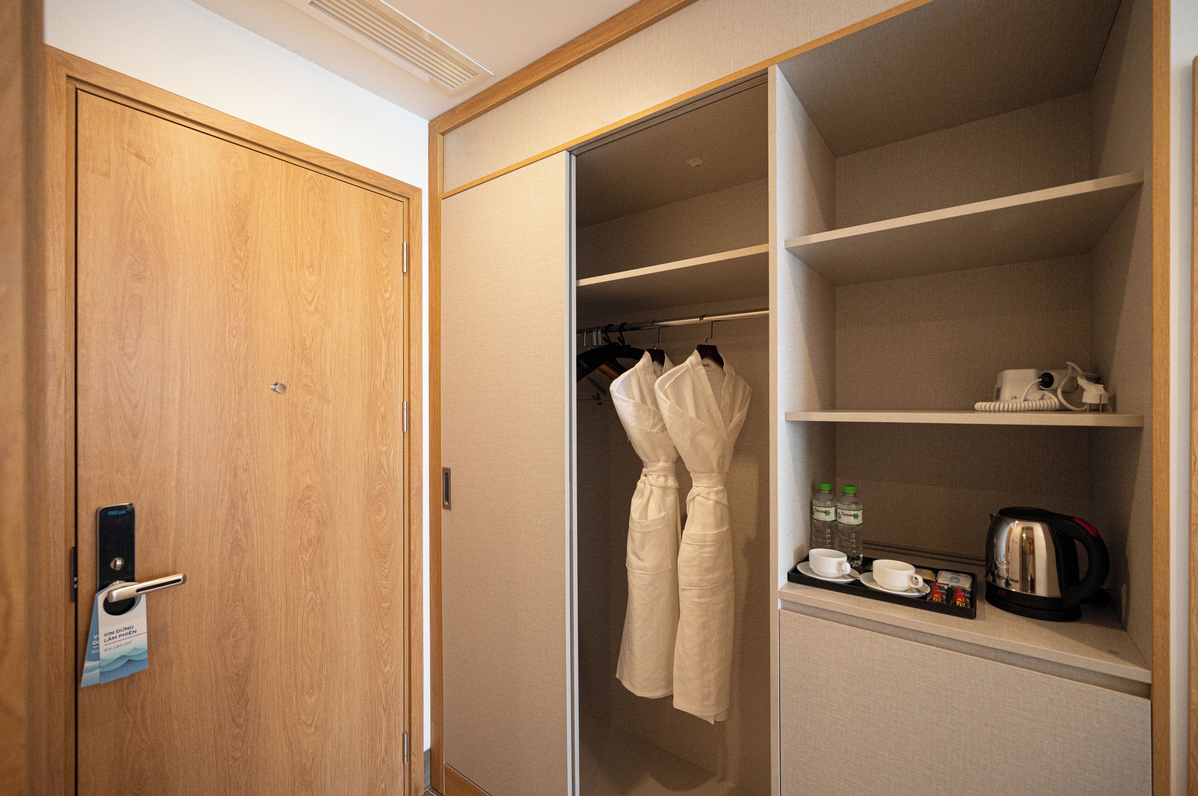 A hotel room with a wooden door, a closet with hanging robes, and a shelf with a kettle, cups, and bottles.