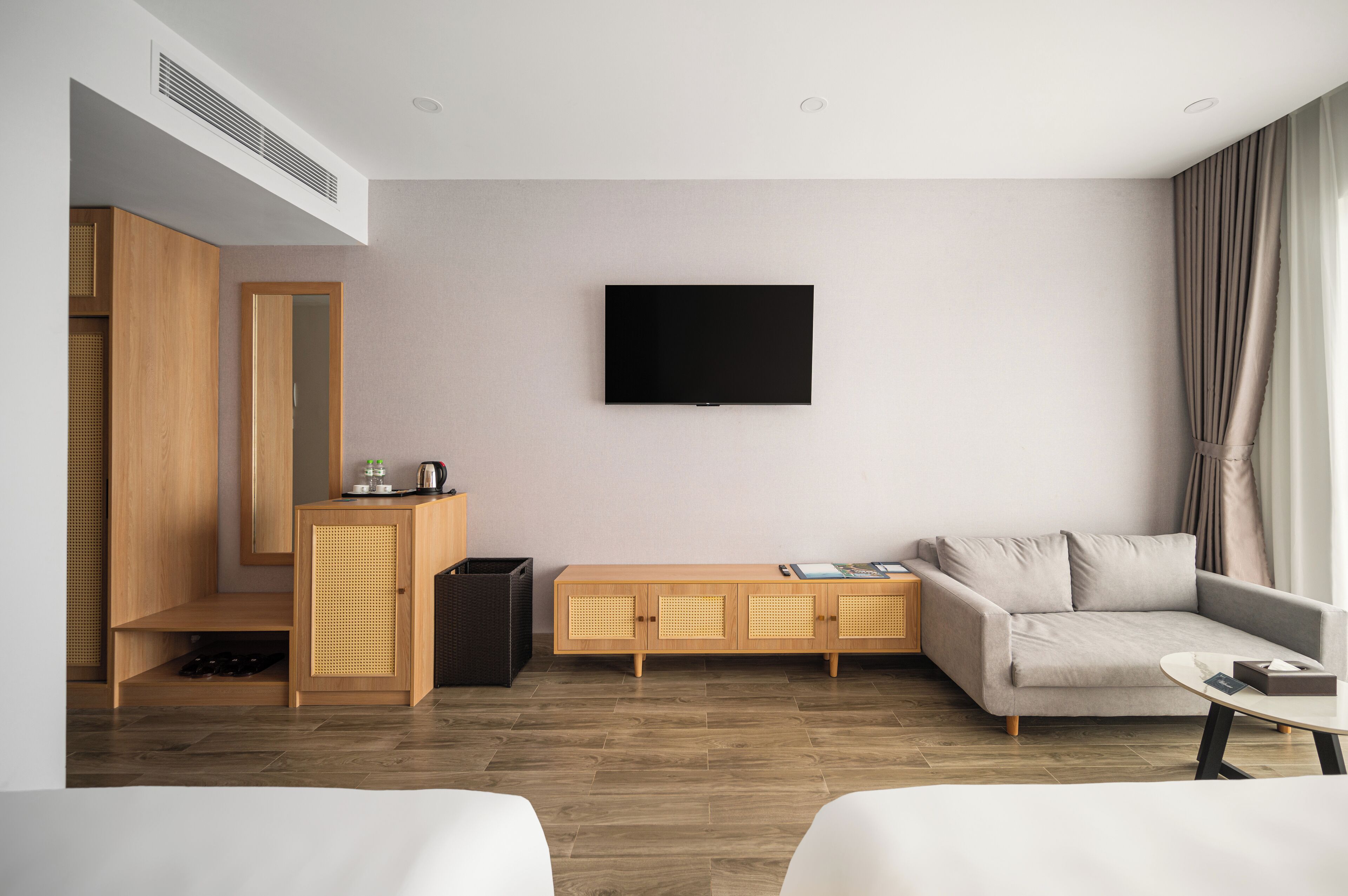 A modern hotel room with a flat-screen TV mounted on the wall, a sofa, a small coffee table, and a wooden cabinet with a wicker basket.