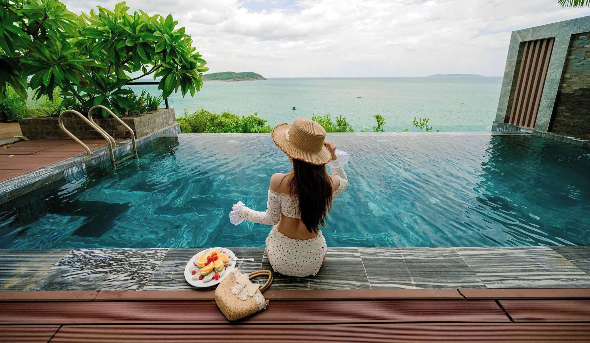 A woman sitting by a pool with a view of the sea, a plate of fruit, and a straw bag.