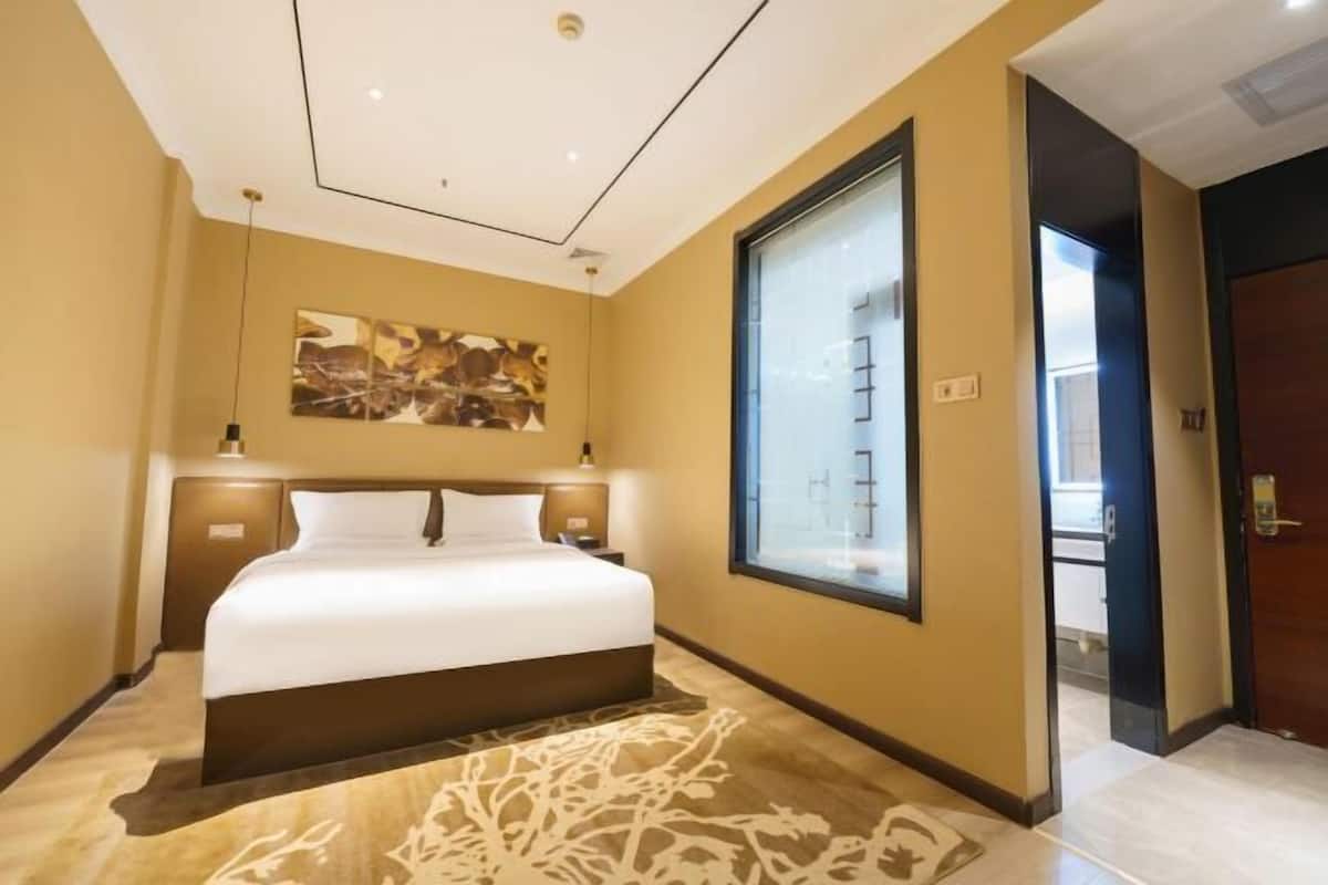 A hotel room with a large bed, a window, a painting on the wall, and a door leading to another room.