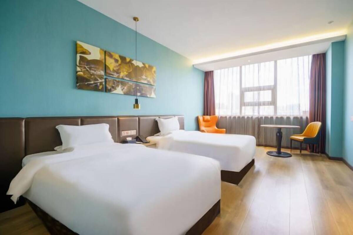 A hotel room with two beds, a table, a chair, and a large window with curtains.