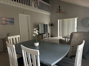 A dining area with a grey table, white chairs, a potted orchid, a television, and a window with blinds.