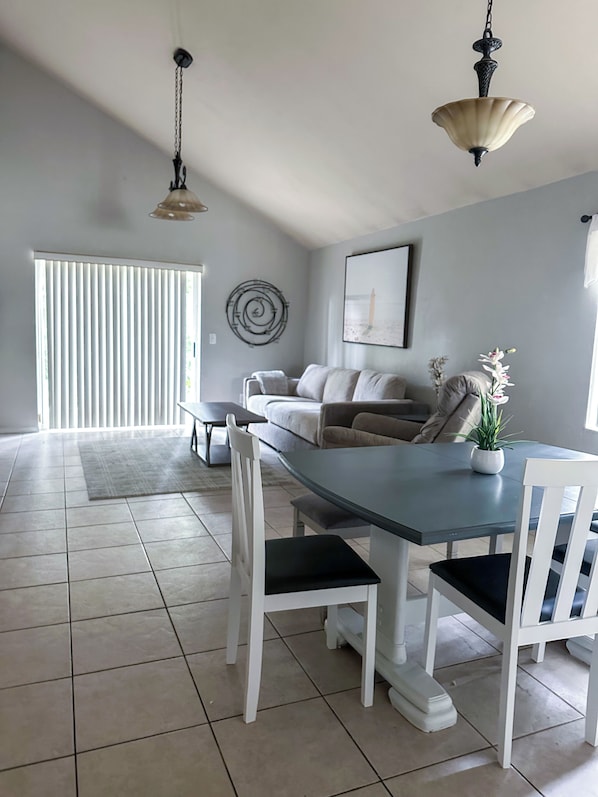 A modern living room with a dining area, featuring a light-colored sofa, a dining table with white chairs, a hanging light fixture, and a large window with blinds.