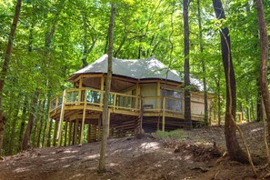 Une cabane perchée dans un arbre, dotée d’une terrasse en bois et d’un étage supérieur abrité, entourée d’une forêt dense.