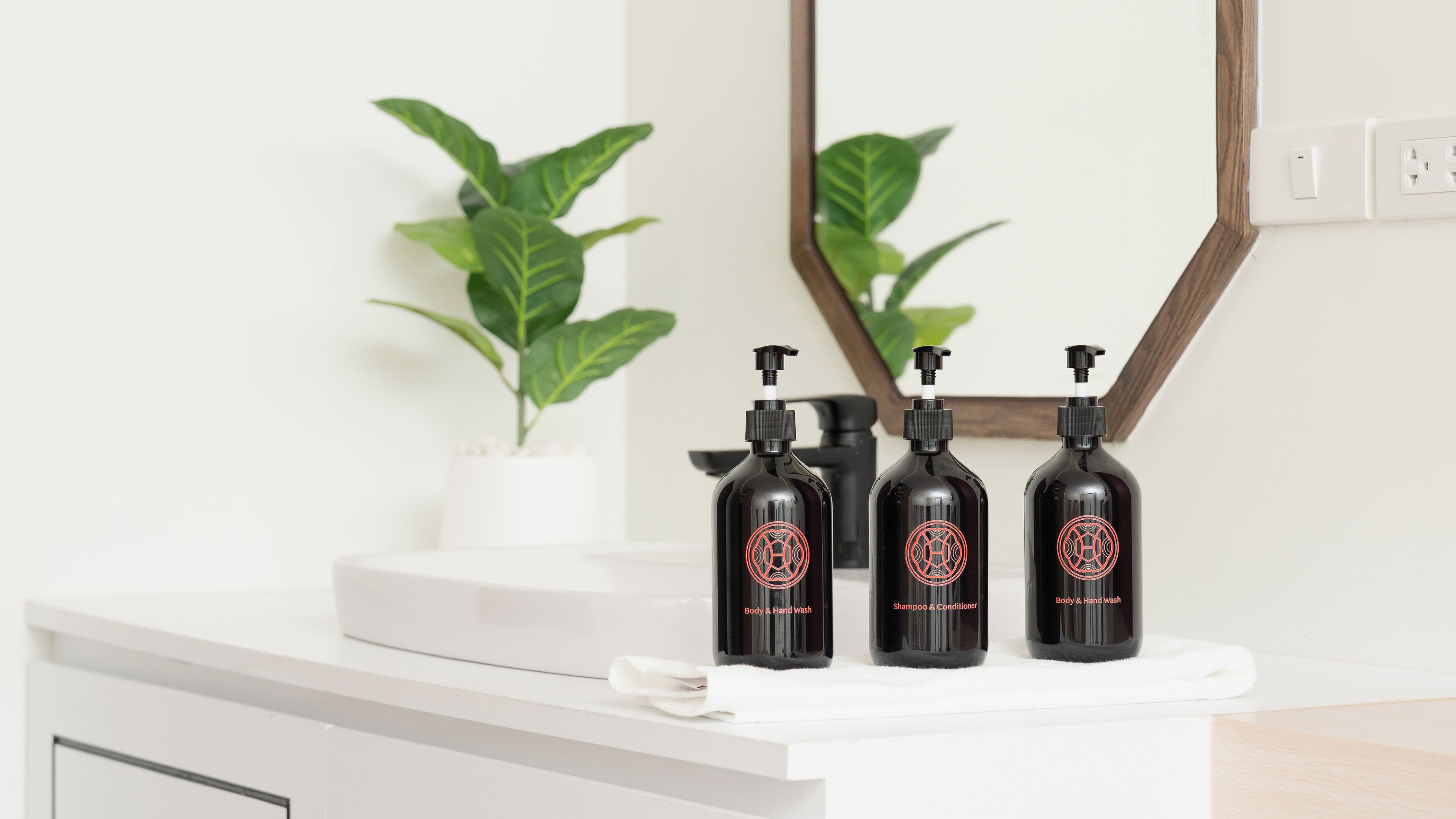 Un mueble de baño con tres botellas negras etiquetadas como “Jabón de Cuerpo y Manos”, “Champú y Acondicionador” y “Jabón de Cuerpo y Manos”. Una toalla blanca está sobre la encimera, y hay una planta en maceta al fondo.
