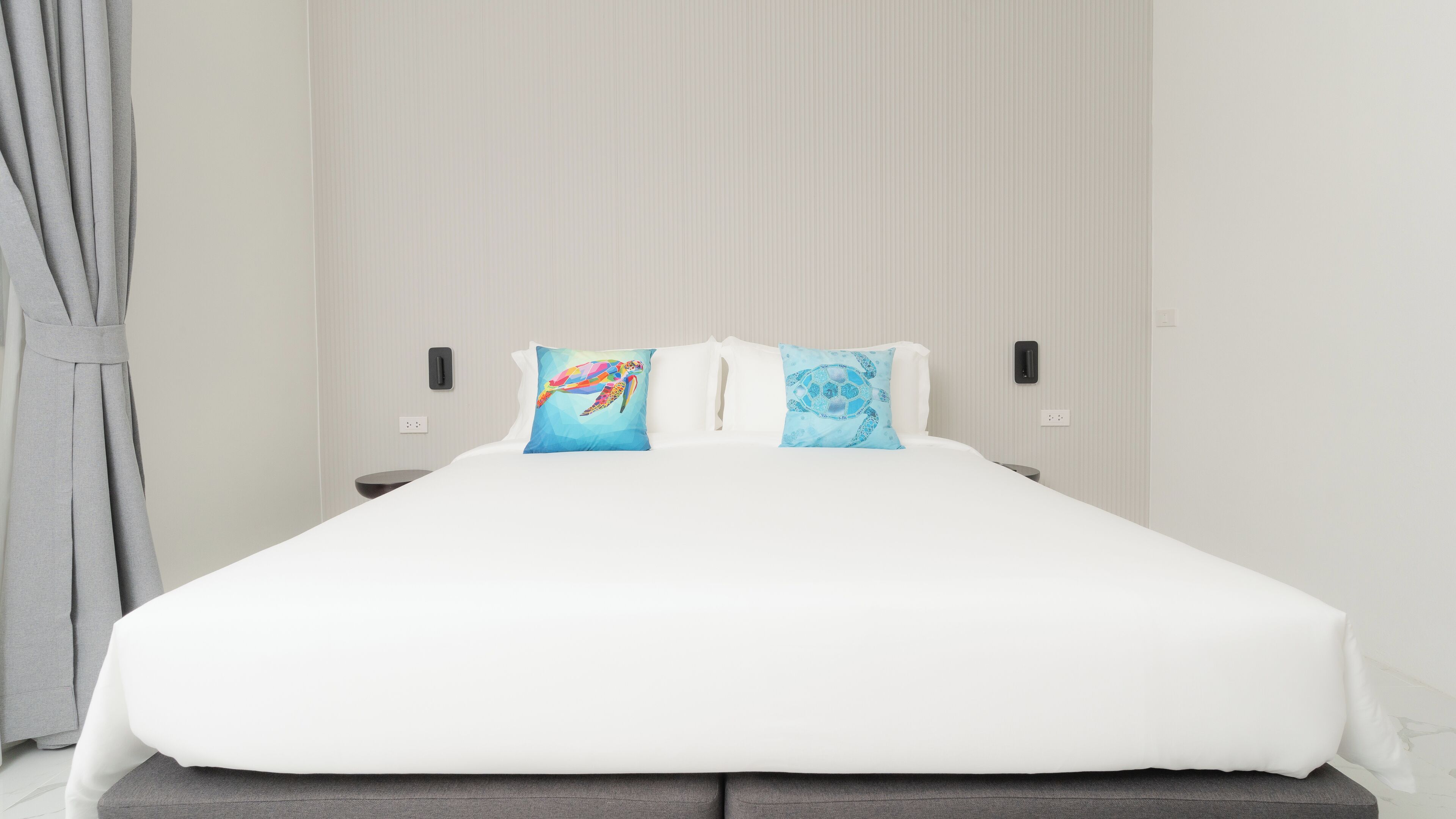 A neatly made bed with white bedding and two colorful pillows, a grey curtain on the left, and a wall-mounted light fixture.