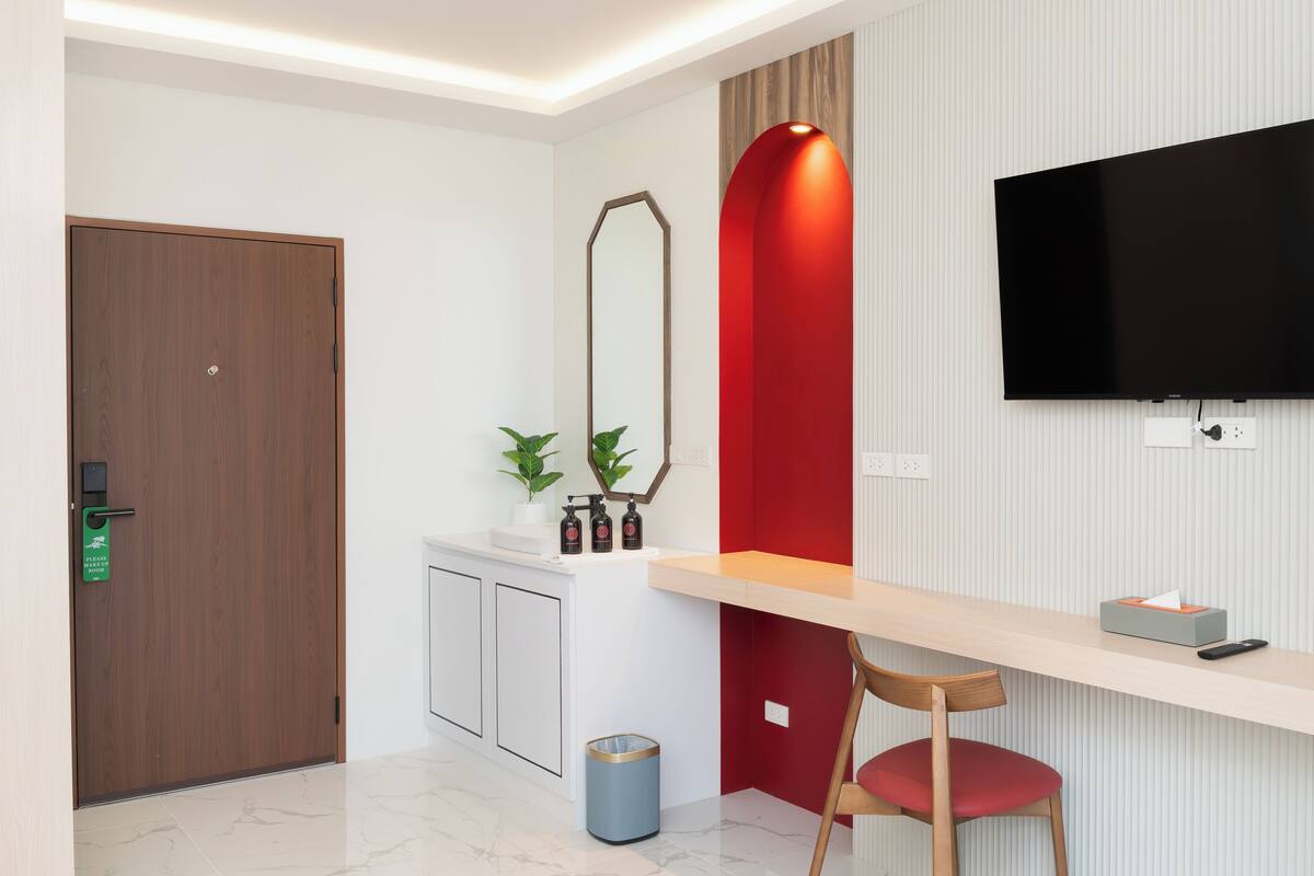 Una habitación de hotel moderna con una puerta de madera, un lavamanos blanco con plantas, una pared de acento roja, un televisor montado y un escritorio con una silla.