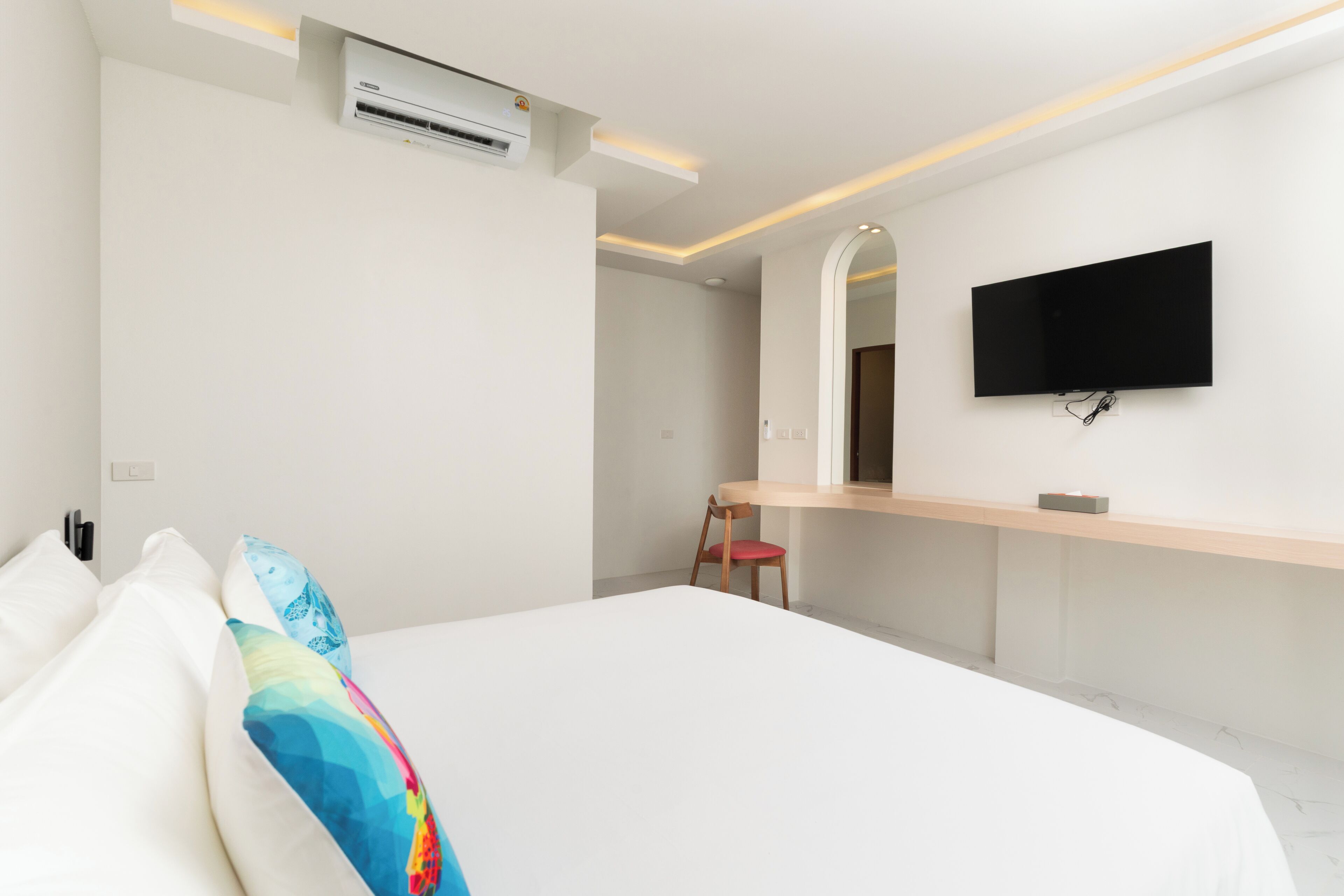 A modern hotel room with a large bed, a flat-screen TV mounted on the wall, a small desk with a chair, and an air conditioning unit.