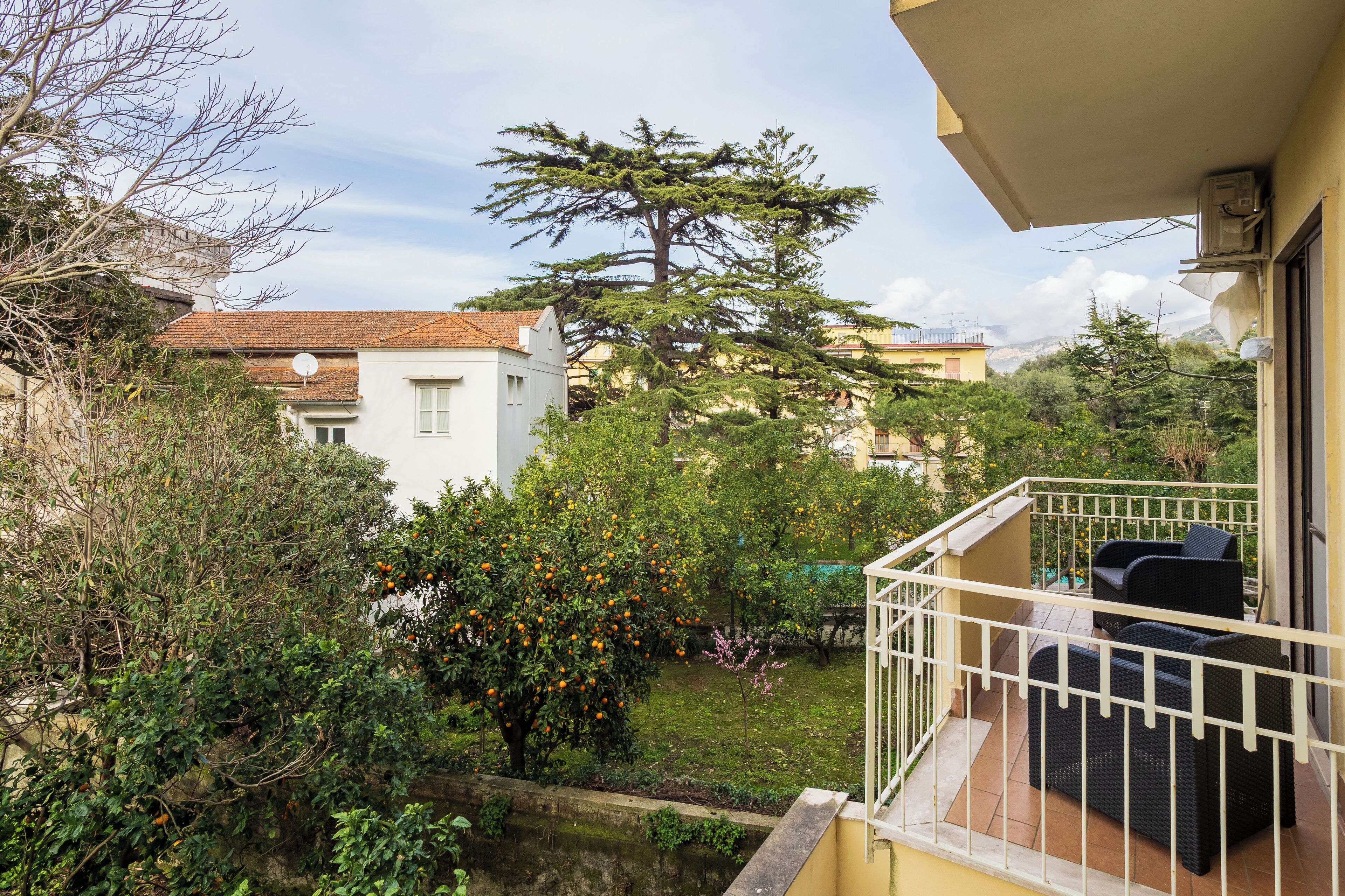 A balcony with outdoor furniture, a garden with trees and shrubs, and a view of neighboring buildings.