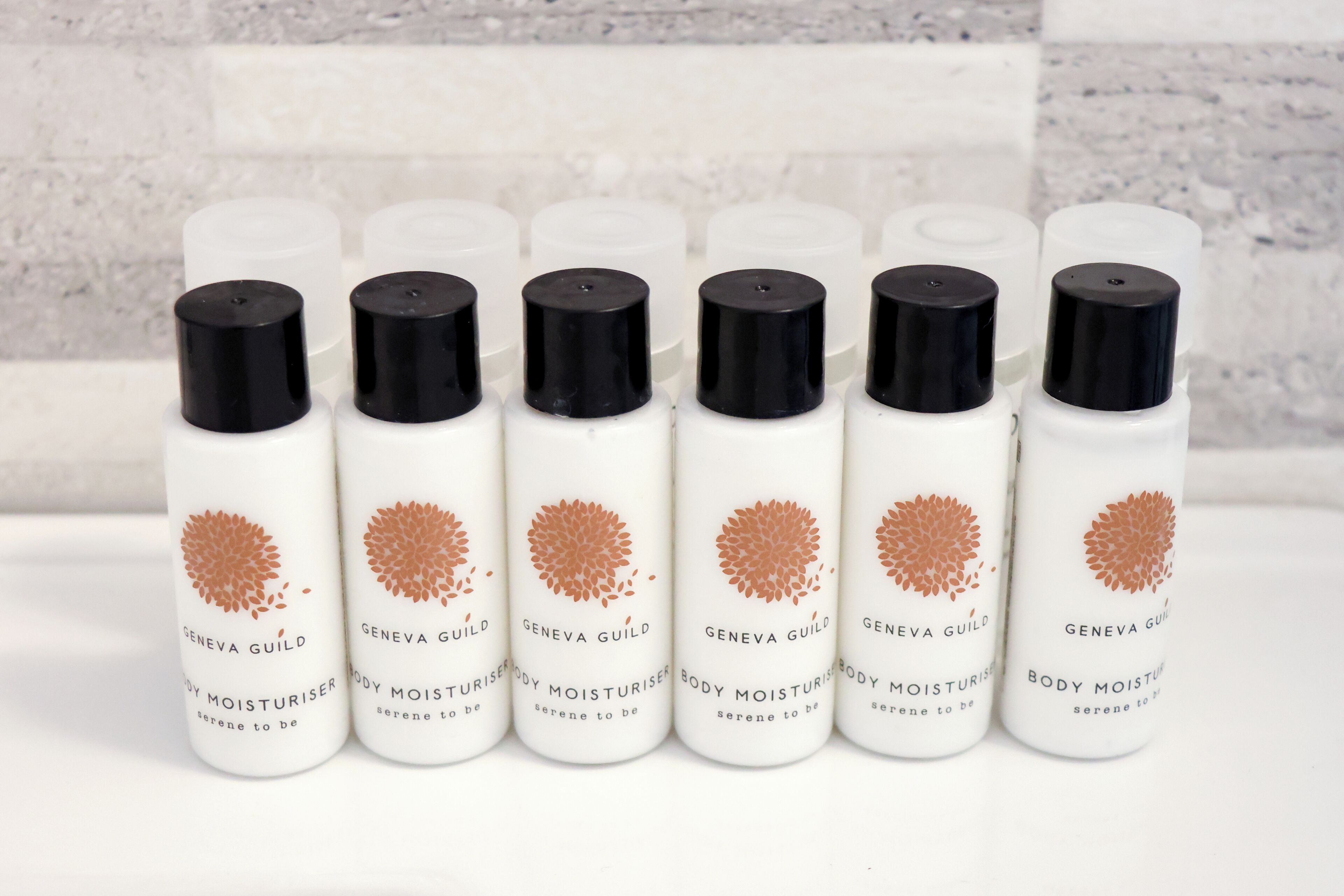 Une rangée de flacons de lotion hydratante pour le corps, dotés de bouchons noirs et d’étiquettes blanches, disposés sur une surface blanche.