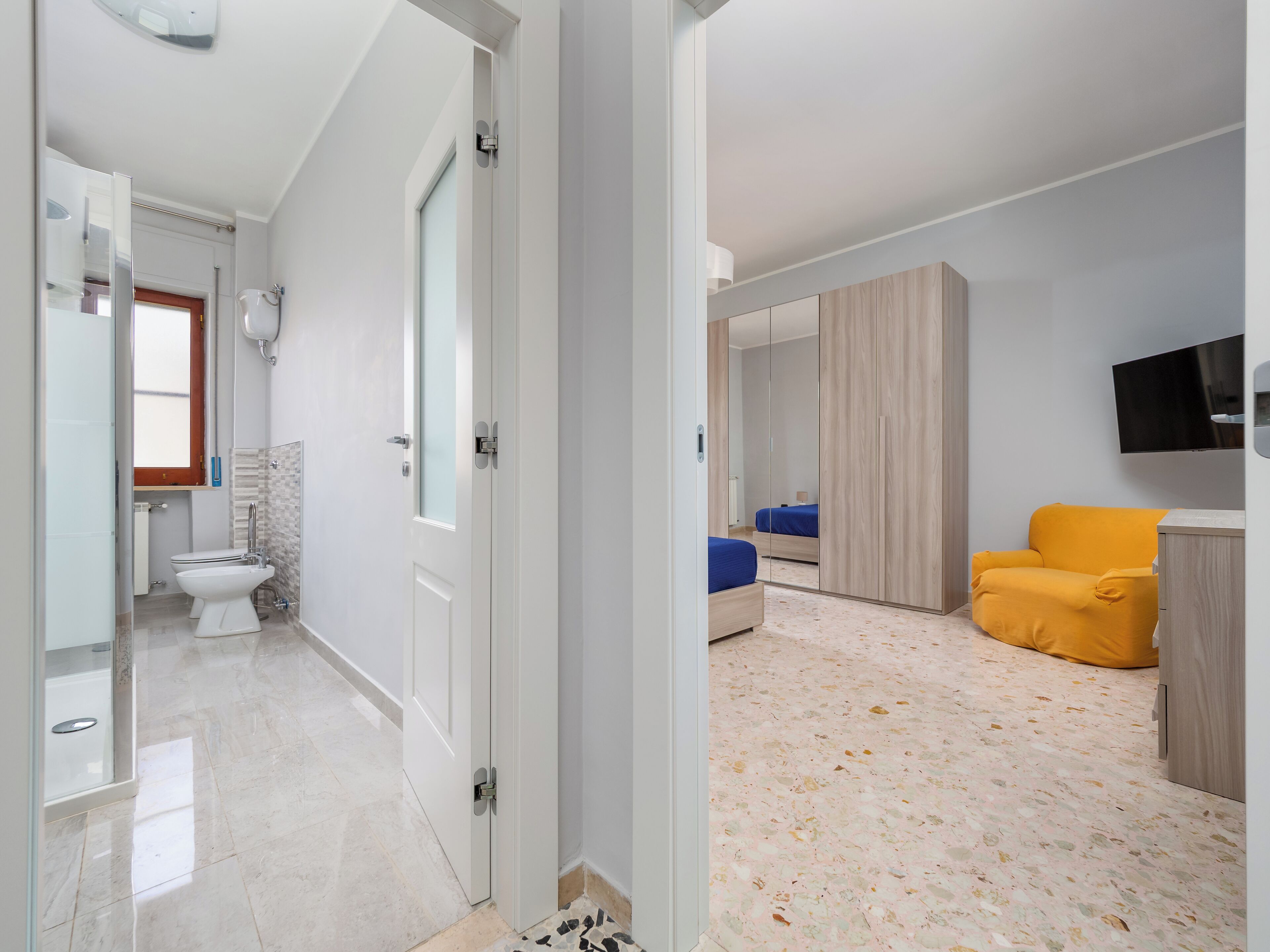 Une chambre d’hôtel moderne, dotée d’une salle de bain, d’un lit avec une couvre-lit bleue, d’un fauteuil jaune et d’un téléviseur fixé au mur.