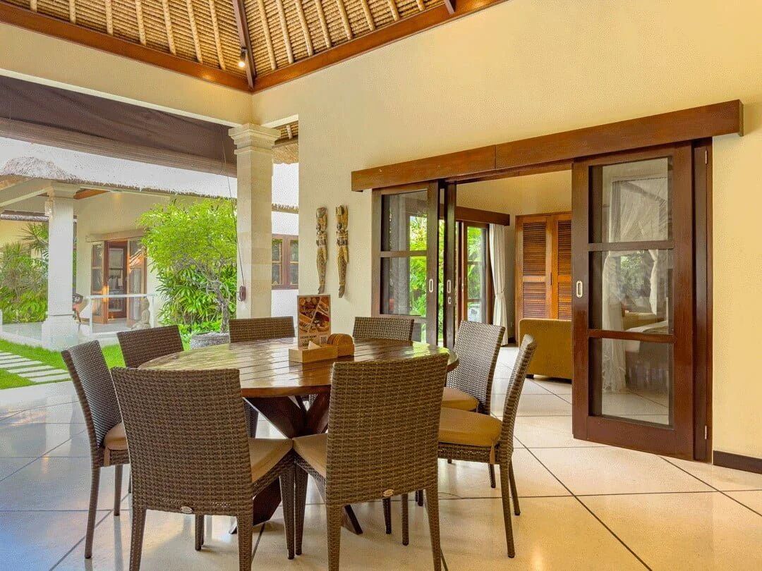 A dining area with a round table and wicker chairs, large windows, and a view of greenery outside.