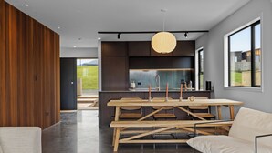 A modern kitchen with a central island, a dining area with a wooden table and chairs, and a large window offering a view of the outdoors. - Studio Āio - Raglan Retreat With Sauna (Raglan)