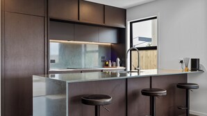 A modern kitchen with a large island, dark wood cabinetry, and a stainless steel range hood. - Studio Āio - Raglan Retreat With Sauna (Raglan)