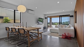 A modern living room with a dining area, large windows, and a view of the outdoors. - Studio Āio - Raglan Retreat With Sauna (Raglan)