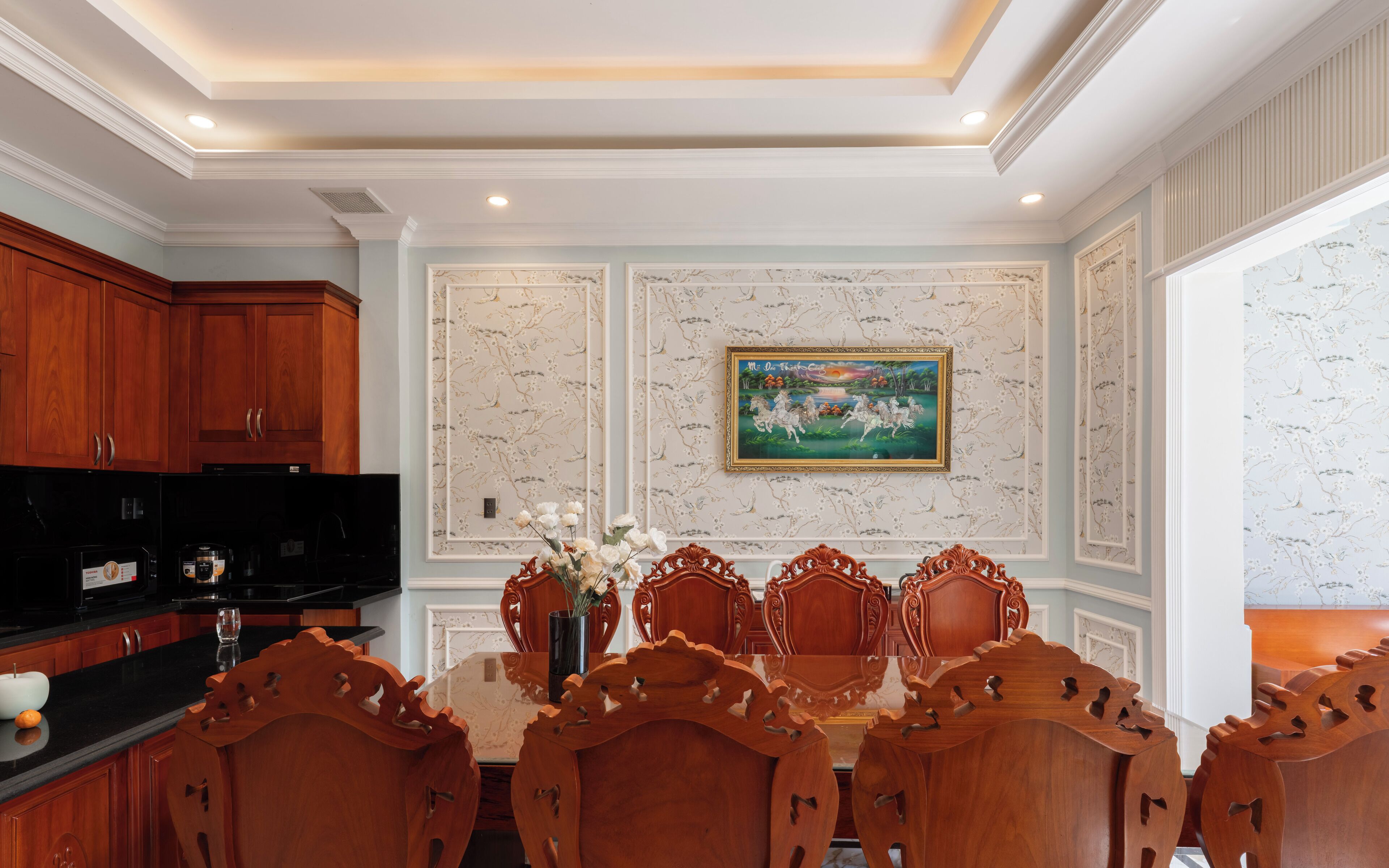 A dining area with a wooden table and chairs, a painting on the wall, and a kitchen with wooden cabinets.