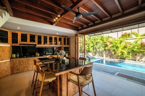 A spacious kitchen with wooden cabinets and a dining area with a glass table. - Island Getaway Villa (Denpasar)