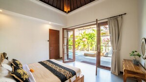 A bedroom with a bed, a wooden nightstand, a door leading to a balcony, and a view of a pool area. - Island Getaway Villa (Denpasar)