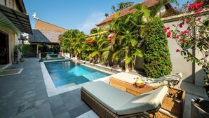 A swimming pool with lounge chairs, surrounded by lush greenery and a building with a tiled roof. - Island Getaway Villa (Denpasar)
