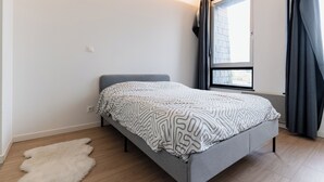 A modern bedroom with a grey bed, a white patterned throw, a white furry rug, and a large window with curtains.