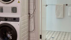 Een gestapelde wasmachine en droger tegen een witte bakstenen muur, met een raam en een deur die leidt naar een betegelde badkamer.