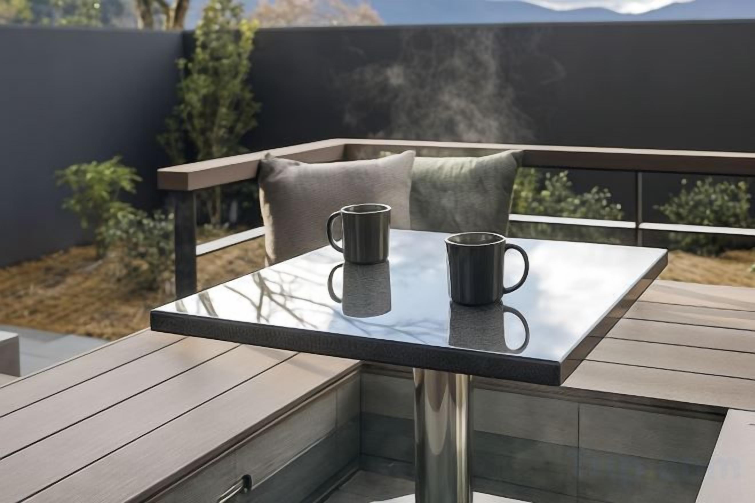 Ein Tisch mit Glasplatte, auf dem zwei Tassen stehen, auf einer Terrasse mit Holzfußboden und Metallgeländer.
