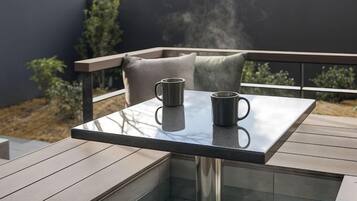 Ein Tisch mit Glasplatte, auf dem zwei Tassen stehen, auf einer Terrasse mit Holzfußboden und Metallgeländer.