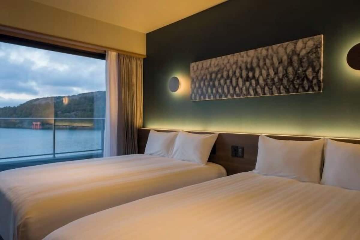A hotel room with two beds, a large window with a view of a body of water and hills, and a wall-mounted artwork.