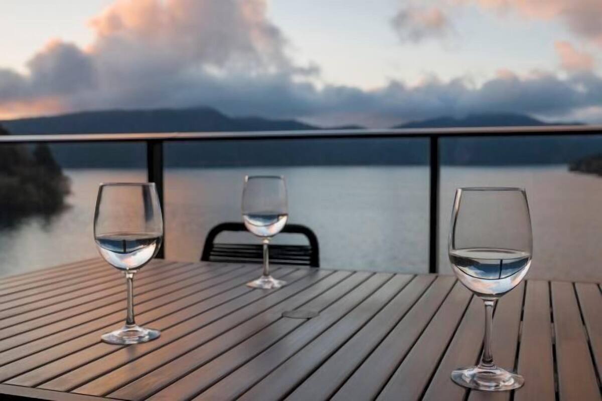 A table with three wine glasses and a chair, overlooking a body of water and mountains.
