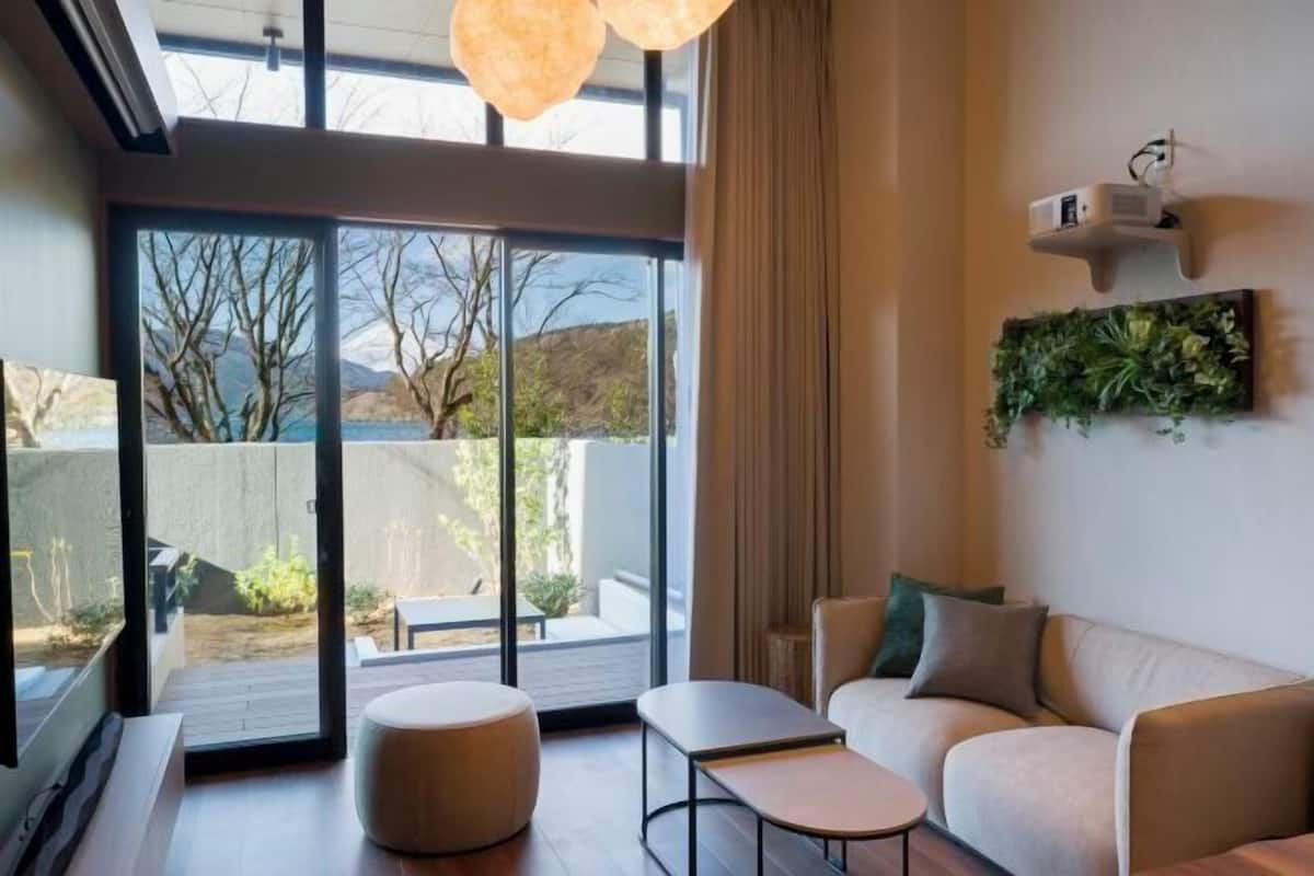 A modern living room with a sofa, a small table, and a round ottoman. There is a large window with a view of the outdoors.