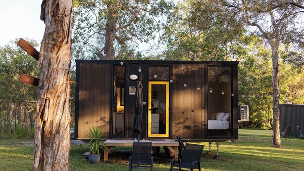 Een moderne cabin met een gele deur, een klein terras en een eethoek met stoelen.