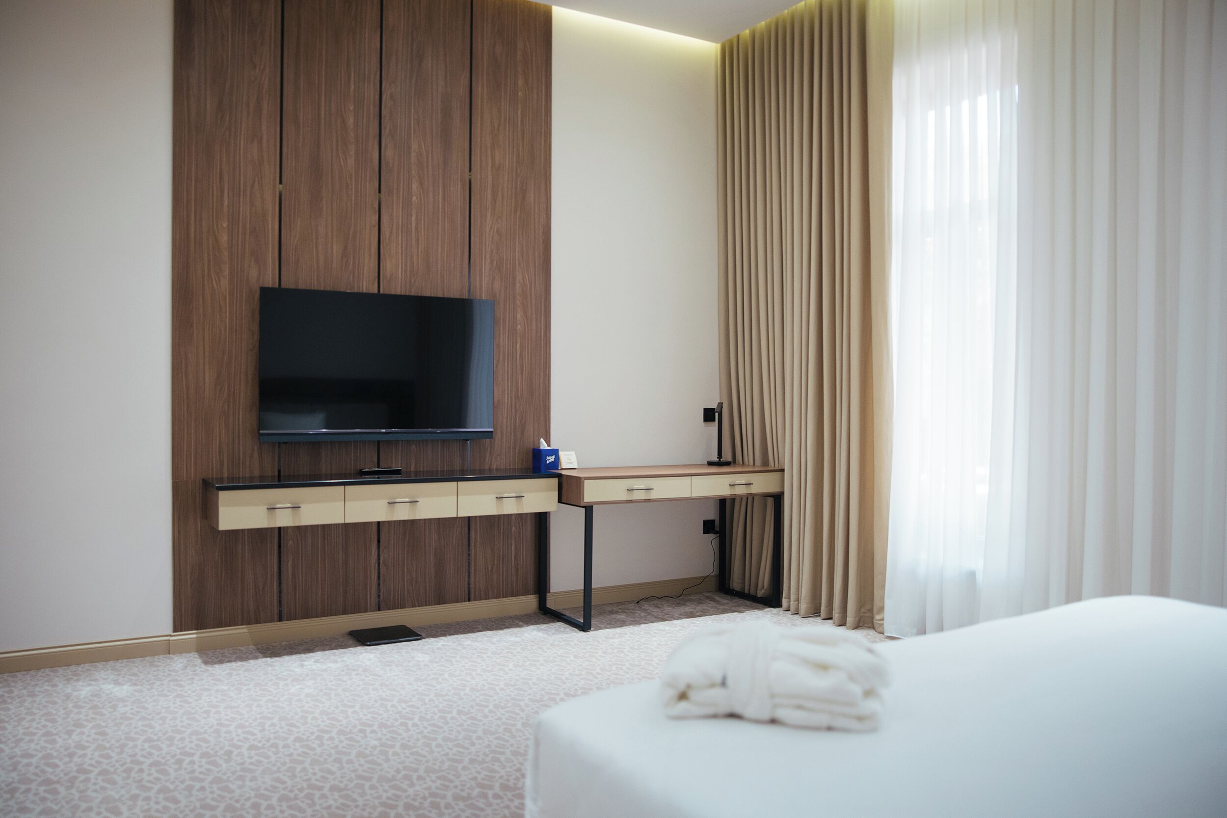 A hotel room with a bed, a desk with a flat-screen TV, a wooden panel wall, and a window with curtains.