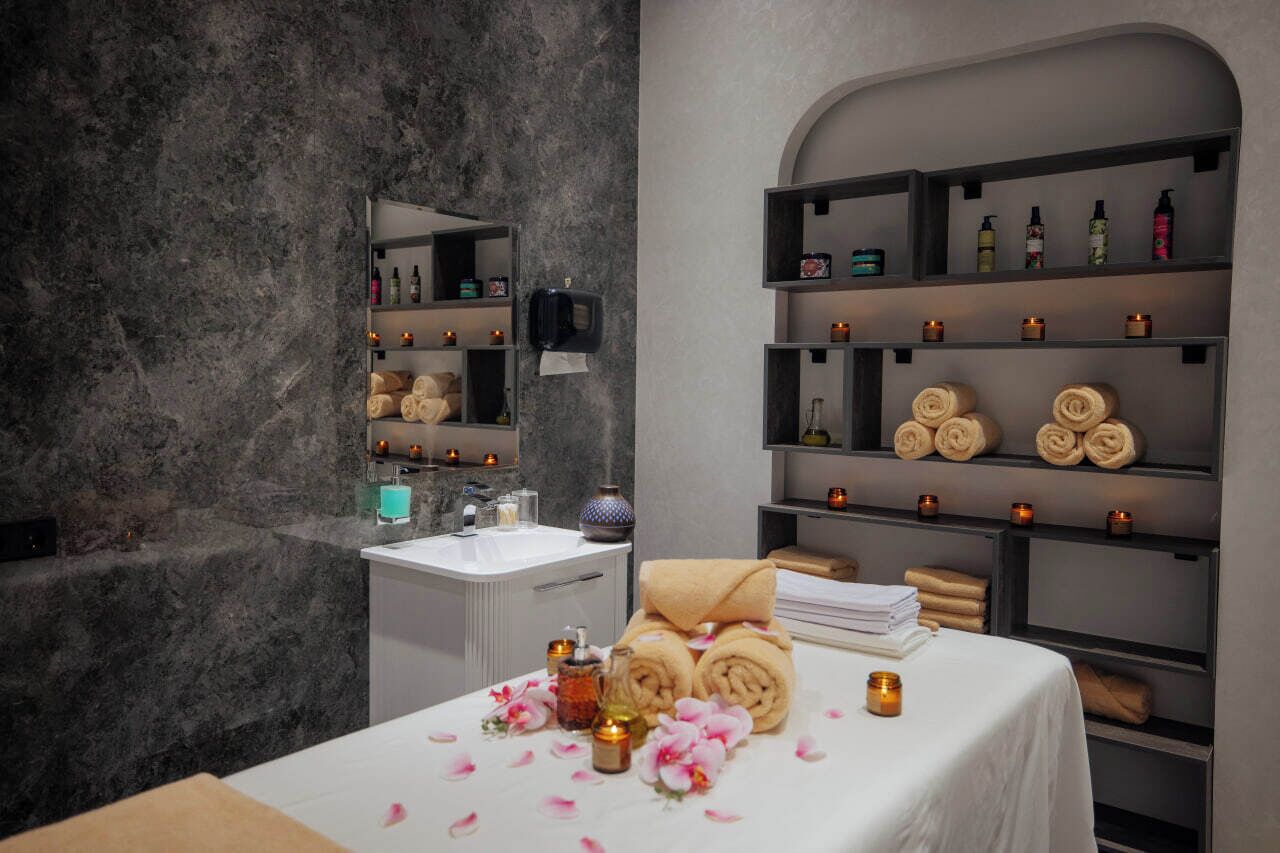 A modern spa room with a marble wall, a white sink, shelves with toiletries, towels, and candles.