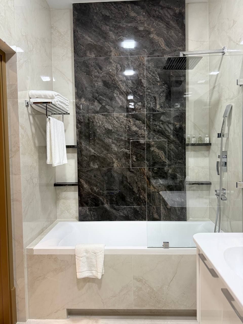 A modern bathroom with a marble wall, a glass-enclosed shower, and a white bathtub.