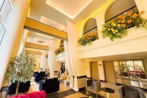 Un hall d’hôtel doté d’un plafond haut, de plusieurs espaces aménagés pour s’asseoir et de plantes décoratives.
