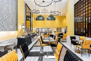 Un restaurant amb una barra llarga, diverses taules i caires. Hi ha llums penjants i una paret amb un disseny decoratiu.
