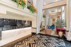 A hotel lobby with a reception desk, a herringbone-patterned floor, a large potted plant, and a view into the room area. - Kastel Bangkok Hotel (Bangkok)